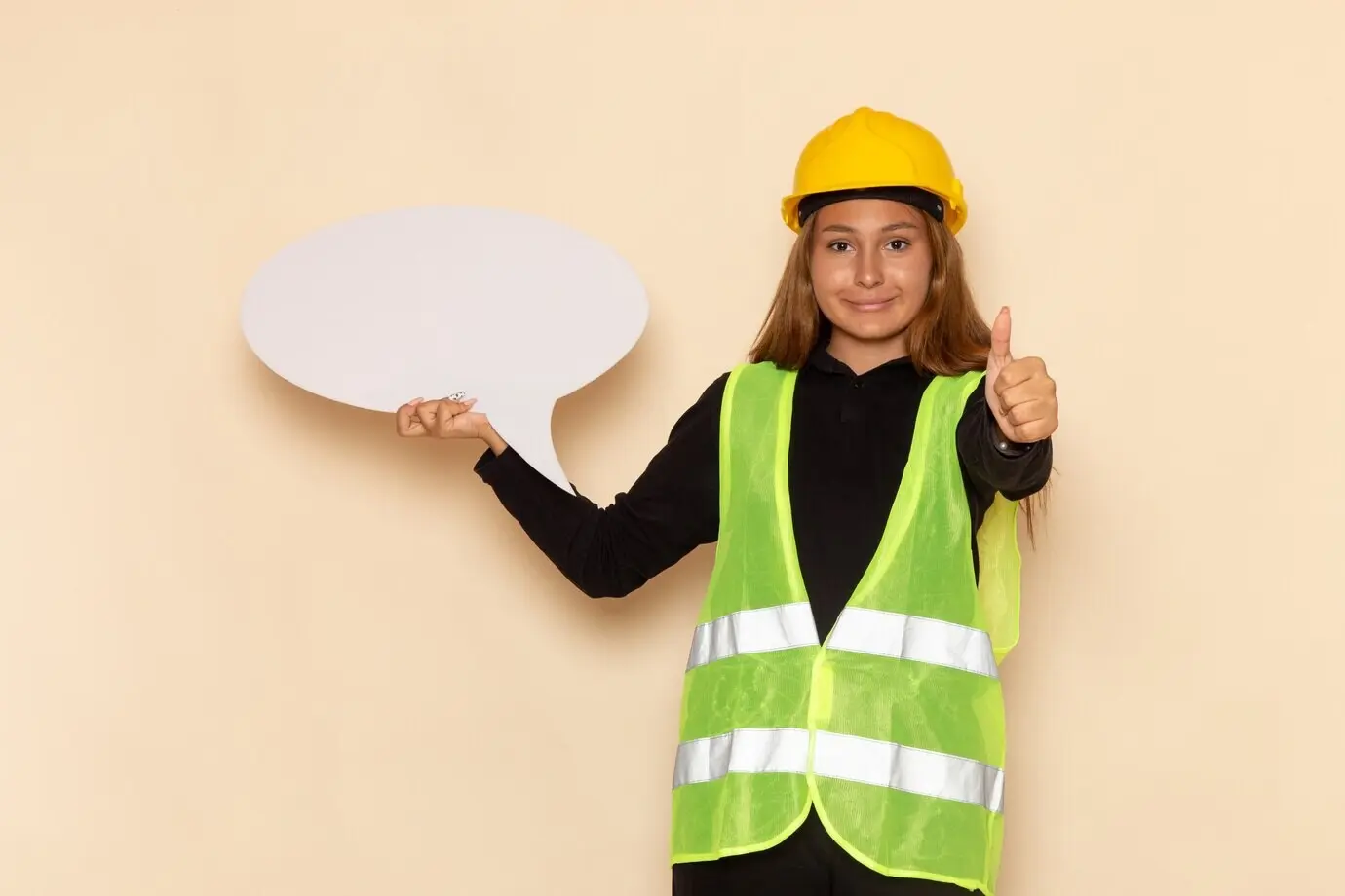 Vista frontal de una mujer constructora y arquitecta con casco amarillo, sosteniendo un cartel blanco y sonriendo, frente a una pared blanca.